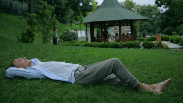 handsome barefoot guy lies down on green park grass to rest and breathes deeply against wooden gazebo slow motion