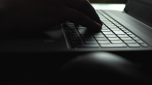 Man working on laptop in dark room / against the light. Close up. Hands typing on a computer keyboard and using computer mouse. Scene A.