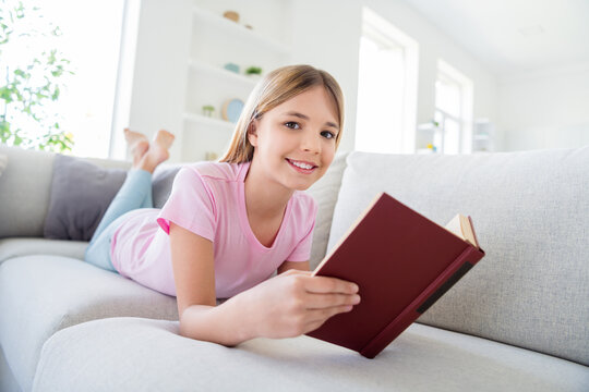 Great remote study concept. Full size photo of positive kid pupil girl lying comfy divan read interesting textbook prepare school project in house indoors