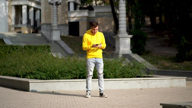 Young man in yellow hoodie puts on headphones and dances, rolling down on curb in park. Adult cheerful male listens to music and enjoys good mood on sunny day