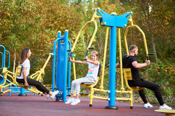 Happy family of three wearing sportswear spending time at the outdoor gym, focus on daughter working out with her father on chest press machine, mother are training legs on background
