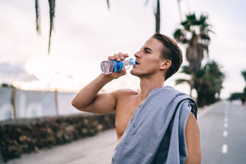 Handsome caucasian male jogger with slim figure drinking water after workout outdoors standing on street, young 20s gyt without t-shirt making pause for refreshment feeling thirsty