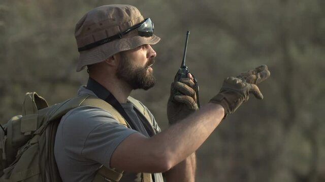 Close-up of bearded man in military clothing, hat and t-shirt, holding backpack and binoculars, explaining route to someone on walkie-talkie, pointing into distance, laughing