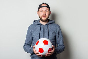 Young man in a cap and hoodie holds a soccer ball on a light background