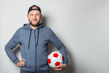 Young man in a cap and hoodie holds a ball in his hands on a light background. Coach