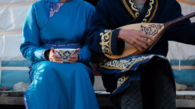 Couple in Traditional Asian Clothes Sitting near Yurt. Man Playing Dombra while his Wife Drinking Hot Tea from a Bowl. Dombra is the Kazakh National Musical Instrument. Slow Motion.