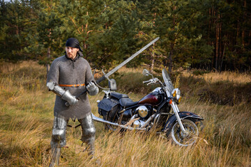 A medieval warrior in armor with a two-handed sword in his hands stands next to a motorcycle...