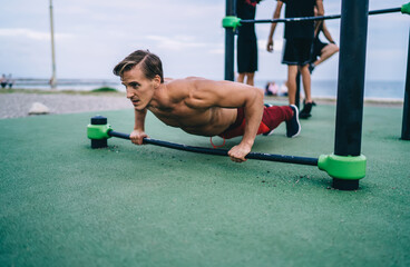 Young Caucasian male with muscular hands pushing up from bars at sports playground using equipment for warming up, sporty shirtless man enjoying actively exercising for keeping perfect body shape