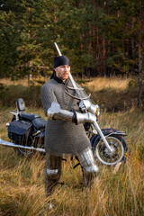 A medieval warrior in armor with a two-handed sword in his hands stands next to a motorcycle...