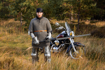 A medieval warrior in armor with a two-handed sword in his hands stands next to a motorcycle against the backdrop of a forest. Knight biker concept.