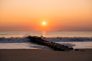 A beautiful sunrise on the last day of Summer at Rehoboth Beach in Delaware. A freight carrying cargo ship is passing by in the distance and the surf is splashing onto the jetty.