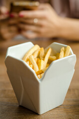 Fries are served at the restaurant in a in white porcelain box styled like take away box. On a wooden table on the background of female hands with a phone on lunch