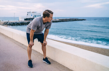 Pensive caucasian male jogger in activewear breathing after cardio workout looking at water in...