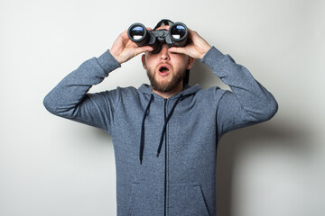 Young man with a surprised face in a hoodie and a cap looks through binoculars on a light background