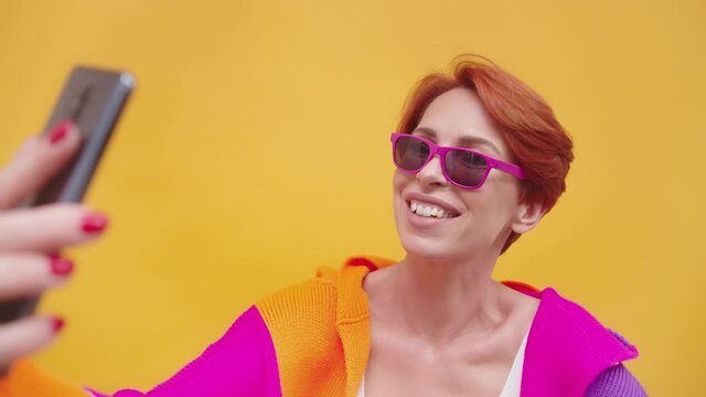 A Lovely Matured Woman In Trendy Eyewear And Colorful Outfit Taking Selfies With Her Smartphone Over Yellow Gold Background - Closeup Shot Slow Motion