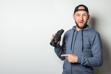 Young man with a surprised face in a hoodie and a cap holds binoculars in his hands on a light background