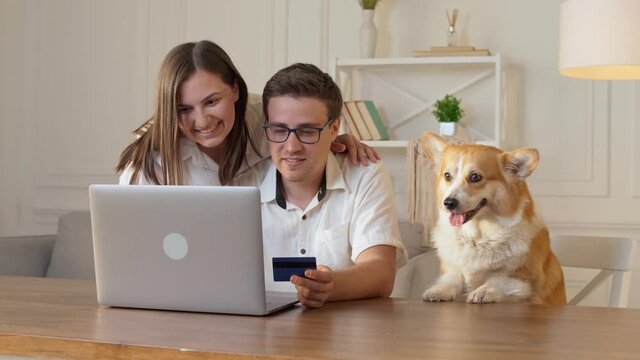 Cheerful Young Couple With a Dog Shopping Online While Sitting Home. Mutual Family's Purchases. Purchase Confirmation by the Internet. Binding a Card for Online Shopping