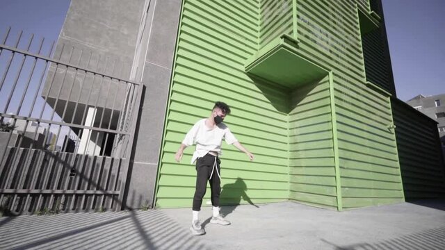 frontal shot traveling movement Caucasian boy curly black hair with short white t-shirt and long black sports pants with gray sports shoes doing parkour and flips free running in building green colors