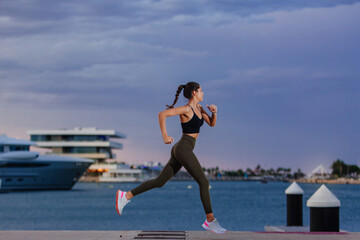 Fototapeta na wymiar Runner dressed in sportswear runs along the sea in a harbor with docked boats. The girl is young and has an athletic body