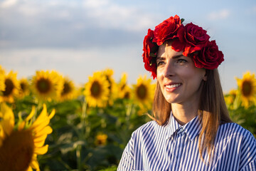 Young woman wearing a wreath of red flowers looks towards the sunset on a sunflower field
