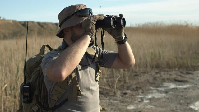 Side view of serious bearded man with backpack, walkie-talkie and watch, wearing military clothing and hat while standing in field, looking through binoculars, hunting or guarding area