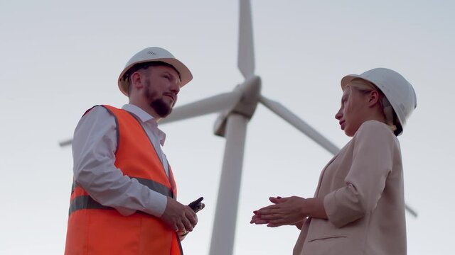 A business woman in a jacket and helmet talking to an engineer a man in an orange vest, a man talking on a radio, against the background of a windmill. Employees of the engineering industry in helmets