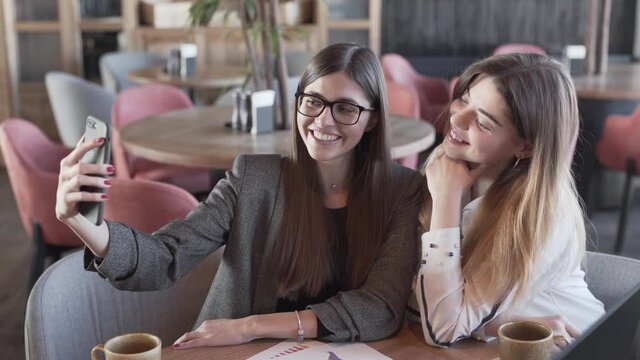 Close-up of two beautiful girls with long hair and magnificent smiles sitting in cafe, working and taking selfies on smartphone.