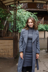 Fototapeta premium young happy smiling pretty girl in gray coat and turtleneck poses outdoors. Street style portrait photo session of attractive female, urban photoshoot of beautiful elegant model
