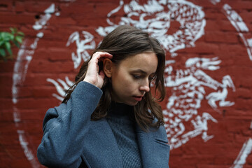 young thoughtful pretty girl in gray coat and turtleneck poses in front of red brick wall with graffiti. Street style portrait of female, urban photoshoot of elegant model