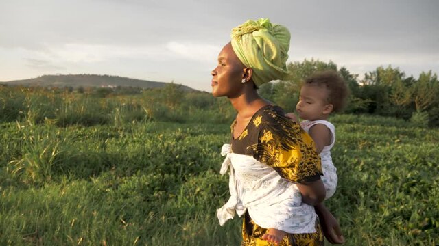 A tracking shot from the side of an African mother with her baby on her back walking by a lake in the golden sunset light.
