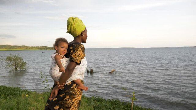 A slow motion shot of an African woman standing by the shores of a windy lake with her baby on her back.