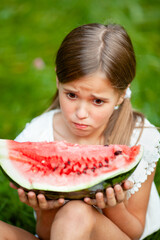 8-year-old girl in white clothes and with two tails on her head eats watermelon on the grass in the park