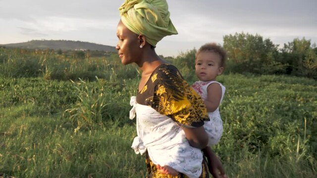 A slow motion side view walking shot of an African mother with a baby on her back walking in the sunset light.
