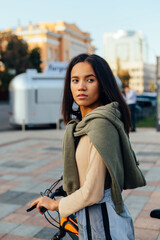 Street portrait of a stylish latin woman in casual clothes with a bicycle standing on the street and looking away. Vertical.