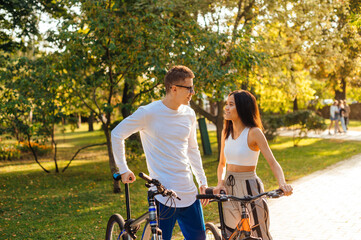 Athletic young people stand with bicycles on the track in the park and communicate with a smile on their faces. Positive young couple walking in autumn park on bicycles.
