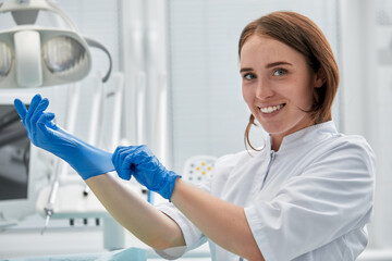 A female dentist puts on gloves against a background of dental equipment in a dental office. Happy patient and dentist concept.