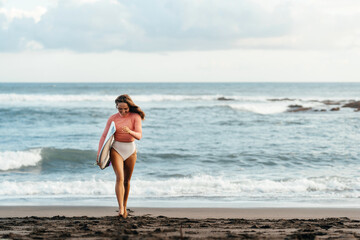 Young attractive surfer woman with white board at sunset on the ocean. Bali Indonesia. Summer time, sports, travel content.