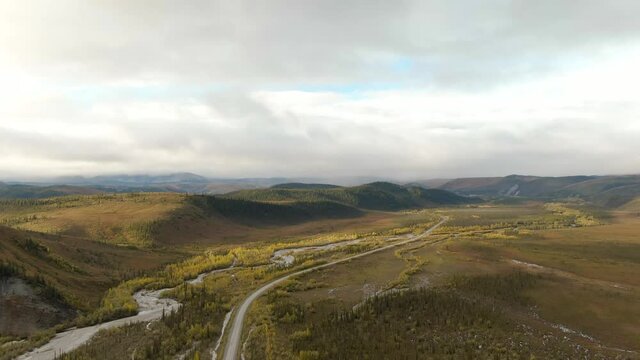 View of Scenic Winding Road surrounded by River, Mountains and Valley in Autumn on a Cloudy Day. Aerial Drone Shot. Taken near Dempster Highway, Yukon, Canada. 4K