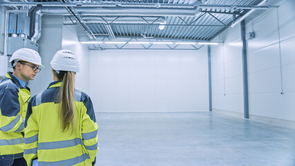 Two Engineers Talking, Standing in the Middle of an Empty Warehouse, Inspecting, Planning Future Factory Layout. Professionals Industrial Designers Strategizing about New Manufacturing Plant Design.