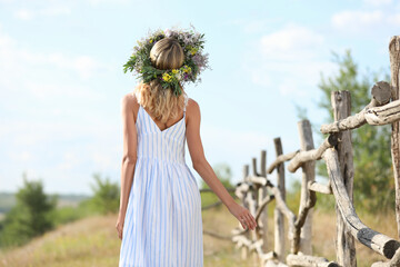 Young woman wearing wreath made of beautiful flowers near wooden fence on sunny day, back view