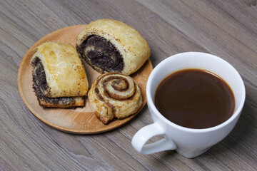 Baked buns stuffed with poppy and cinnamon. Lie on a plate. Nearby is a cup of coffee.