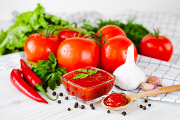 Red sauce or ketchup in a glass bowl and ingredients for his cooking, tomatoes, garlic, basil, parsley, red hot pepper and spices on a wooden white background. Close-up.