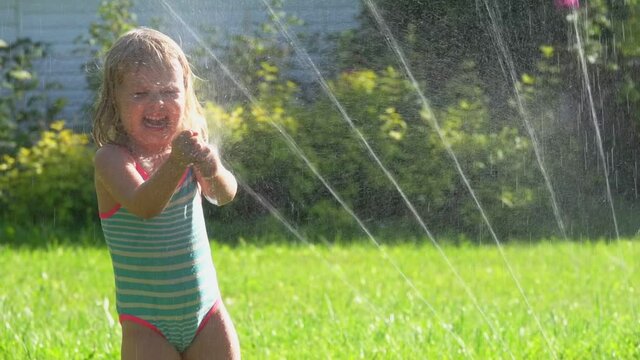The cute little girl in swimsuits is dancing around a revolving water sprinkler on the lawn on the sunny summer day