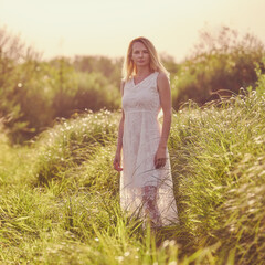 A blonde girl in a white dress walks outdoors in the high field grass