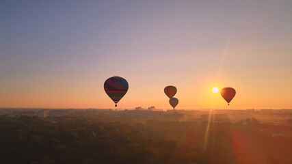 Aerial drone view of colorful hot air balloons flying over green park and river in small european city at summer sunrise