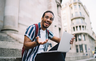 Carefree dark skinned hipster guy satisfied with learning online course outdoors having fun and laughing,positive african american millennial man gesture and looking at camera blogging and share media