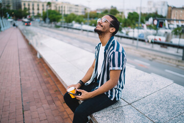 Satisfied young black hipster male with smartphone and takeaway coffee sitting on step in street