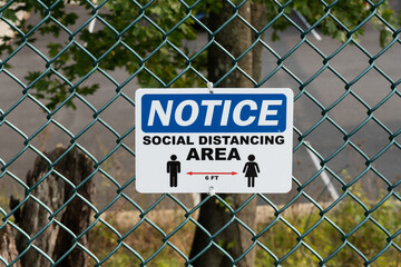 blue and white sign says NOTICE social distancing area with male and female figure and 6 ft apart with a red arrow on a green chain link fence