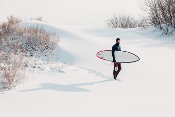 Winter and surfer with surfboard. Snowy beach with surfer in wetsuit.