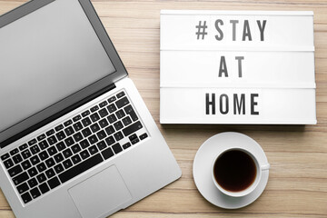 Fototapeta premium Laptop, cup of coffee and lightbox with hashtag STAY AT HOME on wooden background, flat lay. Message to promote self-isolation during COVID‑19 pandemic